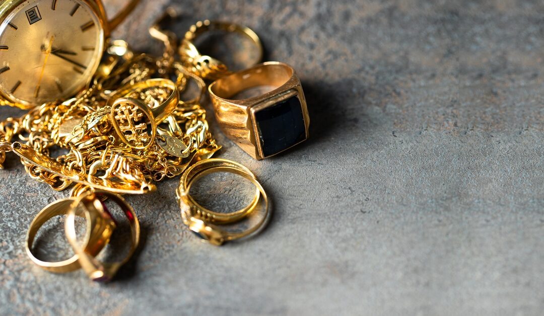 Close-up to multiple gold items, including rings, chains, and a watch, placed on a pile on top of a grey surface.